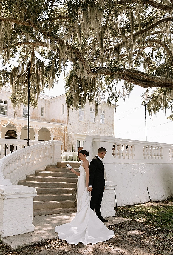 Wedding first look as bride and groom hold hands back to back on a white staircase, bride reads vows under string lights in dappled shade