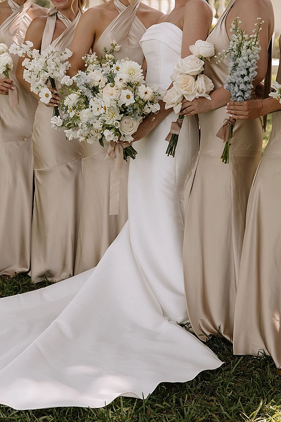 Bridesmaid bouquets of white wildflower and daisy blooms with greenery stems, held beside champagne satin dresses on a sunlit lawn