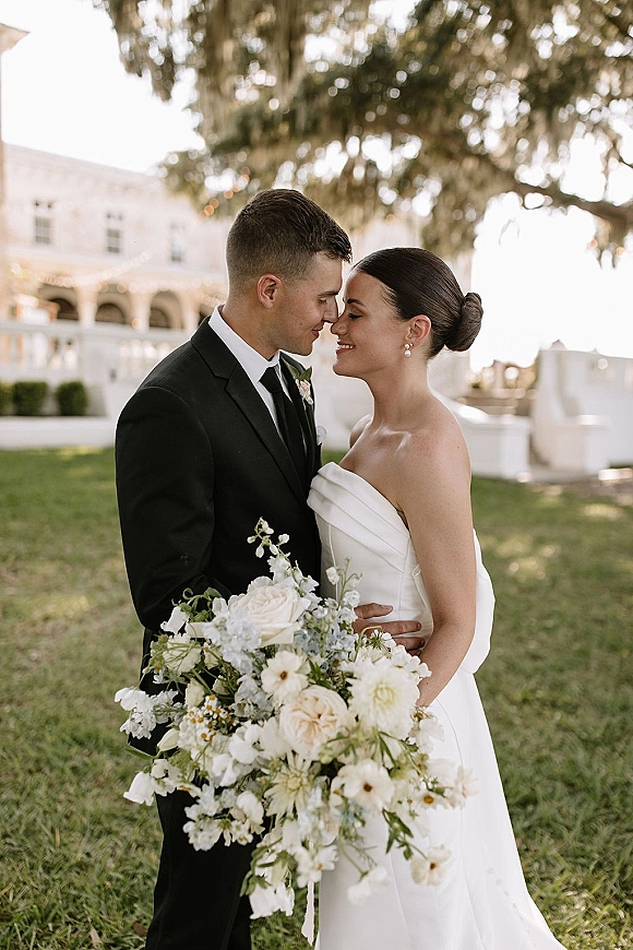 Couple portrait of bride and groom nose kiss, newlyweds embracing on a lawn under mossy tree with string lights by waterfront railing