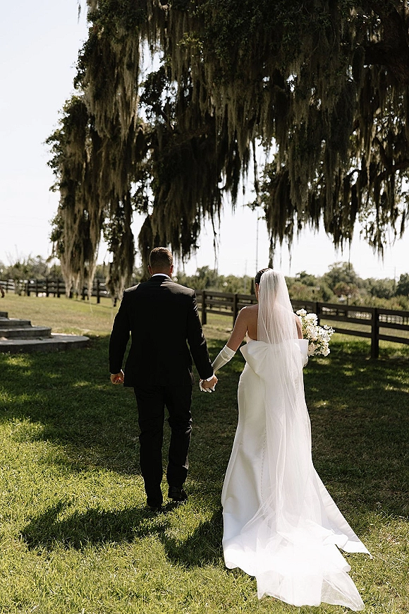 Bride and groom walking hand in hand with cathedral veil and bouquet, seen from behind on a lawn under oak trees with moss