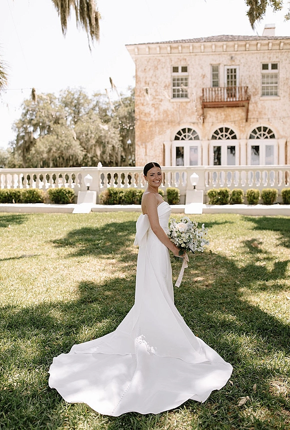 Bridal portrait of a bride holding bouquet in a strapless gown with long train on a lawn by a historic building with arched windows