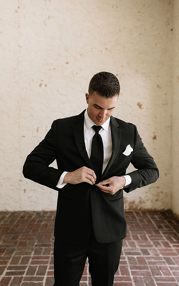 Groom portrait in a black tuxedo, black tie, and pocket square, standing on a brick floor against a textured stucco wall