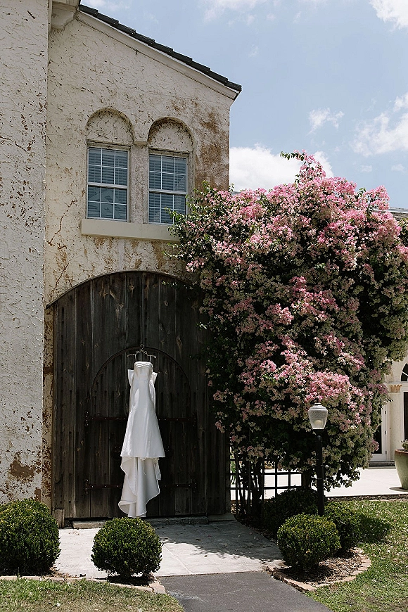 Wedding dress display with a strapless satin gown on a hanger against wooden barn doors, framed by a pink flowering tree and lantern