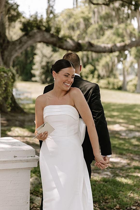First look moment as bride and groom stand back to back holding hands, bride in strapless wedding dress under oak trees on a lawn