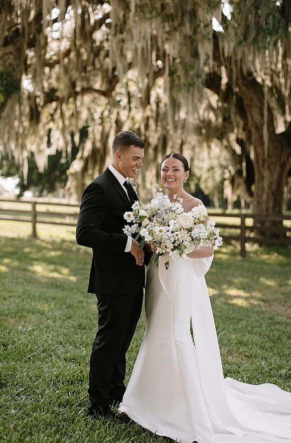 Couple portrait of newlyweds holding bouquet, bride in strapless satin dress beside groom in black tuxedo under oak trees with Spanish moss