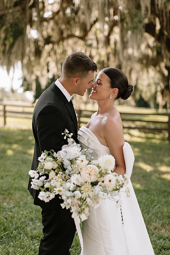 Couple portrait of bride and groom nose kiss, holding a white bouquet on a grassy lawn under oak trees with hanging moss