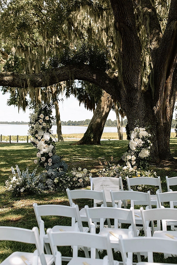 Ceremony setup for an outdoor wedding ceremony with white folding chairs, white and green florals on the ground under oak trees by a lake