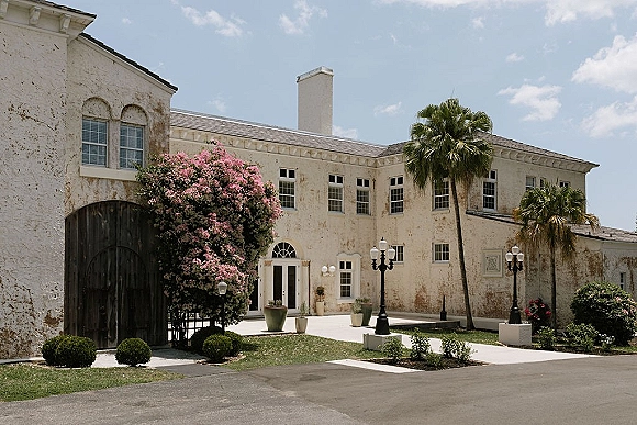 Wedding venue exterior with arched wooden doors and potted plants along a stucco courtyard walkway under palm trees and blue sky