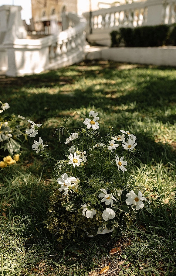 Wedding floral arrangement with cosmos wedding flowers and greenery in a low cluster on grass beside a stone balustrade in a sunlit garden