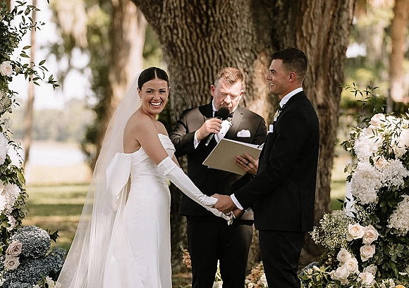 Wedding vows during an outdoor wedding ceremony as bride and groom hold hands by a large tree, surrounded by white rose and hydrangea florals