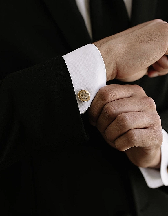 Groom cufflinks as the groom adjusts cufflinks, gold accents on white French cuffs beneath a black tuxedo jacket against a dark background