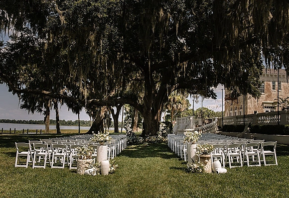Ceremony setup with white folding chairs and wedding aisle flowers under an oak tree draped in moss, with string lights by the waterfront
