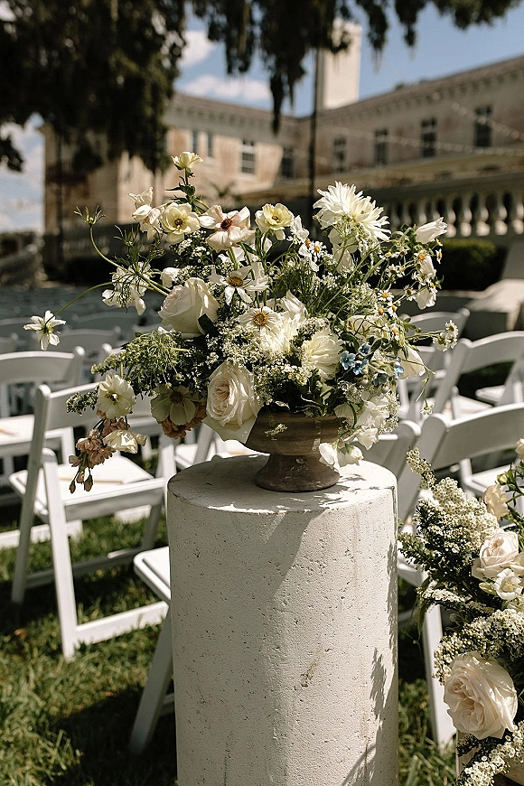 Ceremony floral arrangement of white garden roses, daisies, and wildflowers with greenery in a stone urn beside white folding chairs on a lawn