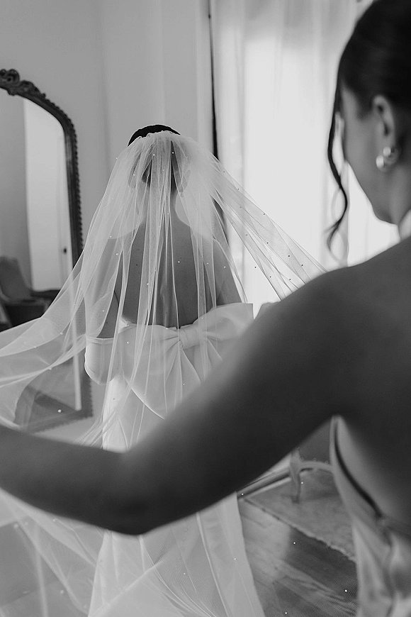 Bridal veil moment as a bride getting ready while a bridesmaid adjusts her cathedral veil, highlighting the bow-back strapless dress by a vanity mirror in window light