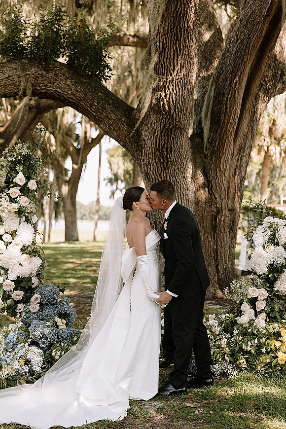 Wedding kiss portrait of bride and groom kissing under a rose and hydrangea floral arch, long veil and gloves, lakeside oak with hanging moss behind