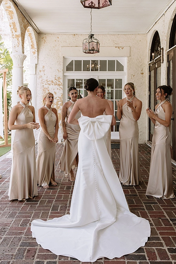 Bridesmaid reveal as bridesmaids react to the bride’s oversized back bow gown and long train on a covered porch with lanterns