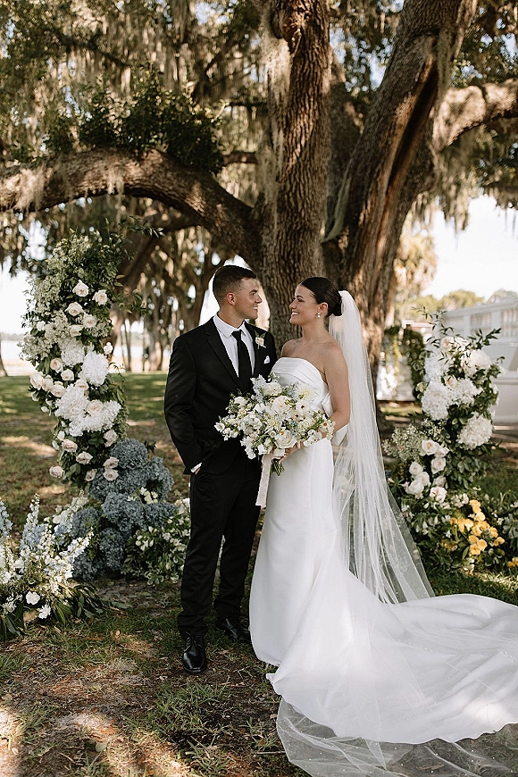 Couple portrait in an outdoor wedding portrait, bride in strapless dress and long veil beside groom in black tux under mossy oaks