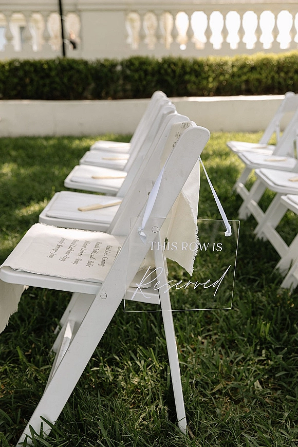 Ceremony reserved sign on a clear acrylic reserved sign tied with a white ribbon to a folding chair, programs on seats on a lawn