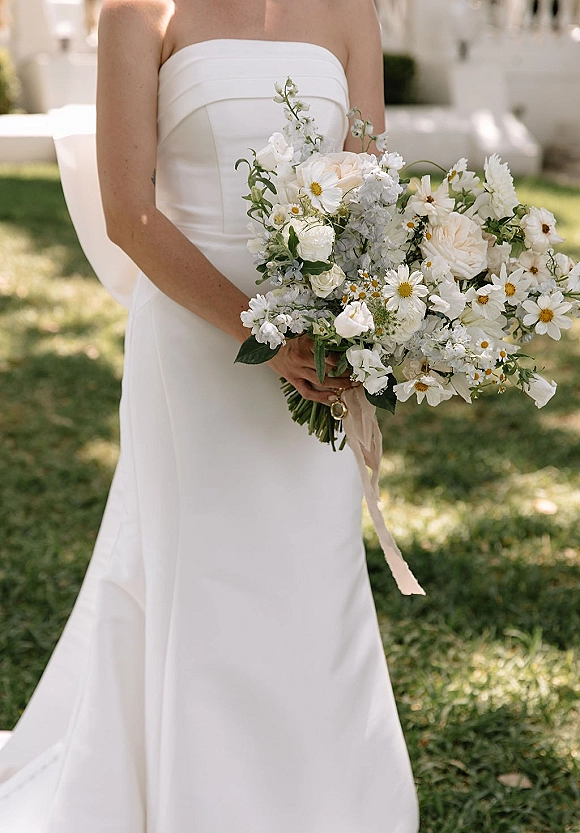 Bridal bouquet of daisies and roses, a white wedding bouquet with greenery and ribbon wrap held against a strapless dress outdoors