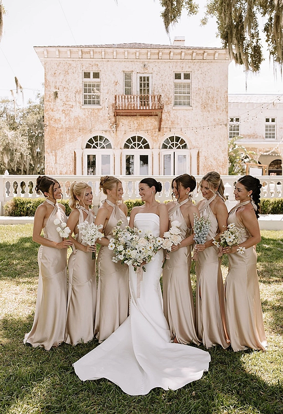 Bridesmaids portrait with bride with bridesmaids in neutral satin dresses holding white-and-green bouquets before a historic facade with arched windows