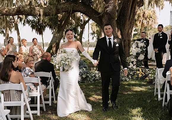 Wedding recessional as bride and groom walk hand in hand down the aisle, bride laughing with bouquet beneath oak tree and moss by lake