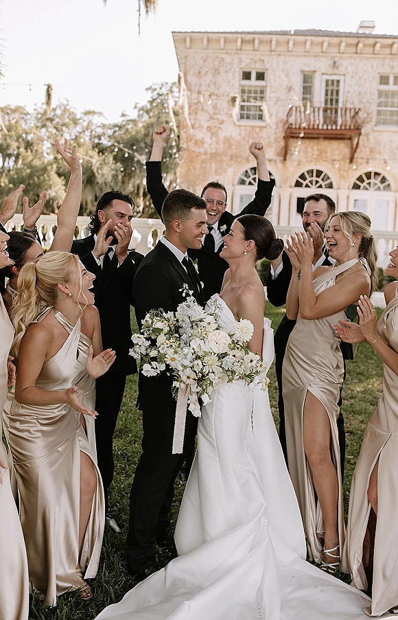 Wedding party photo of bride and groom with bridesmaids cheering, bride holding a white bouquet with greenery on a lawn before a historic facade with string lights
