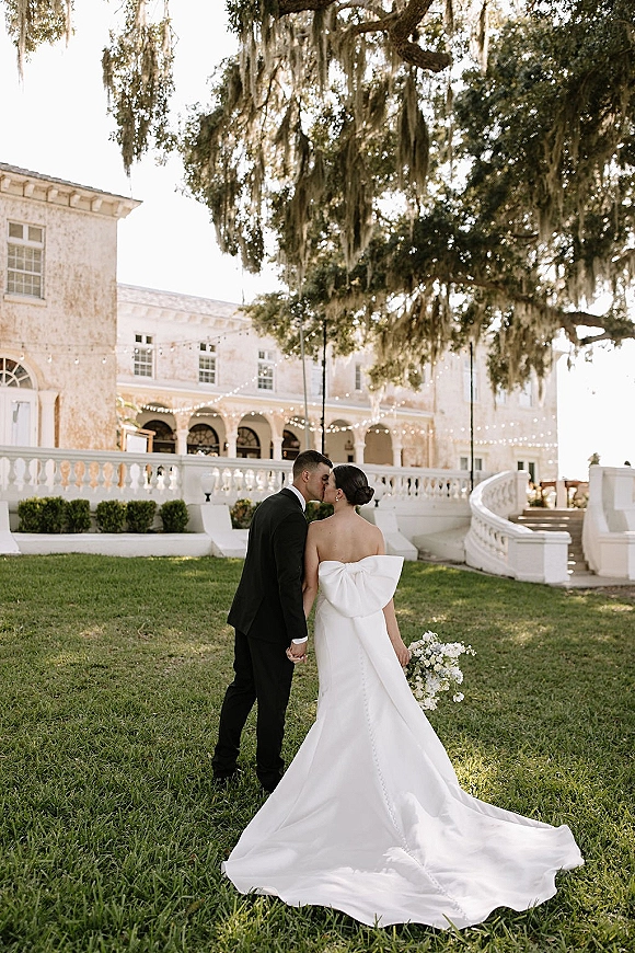 Wedding kiss portrait of bride and groom kissing under string lights on a manicured lawn by a stone veranda with mossy oak tree