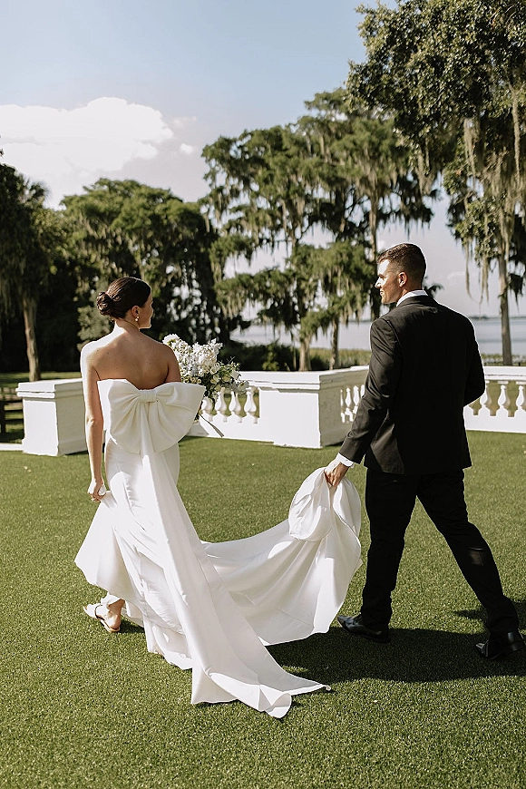 Couple portrait of bride and groom walking away, bride holding bouquet as groom lifts her dress train on a lawn by mossy trees and water