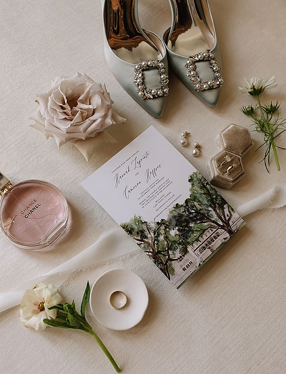 Invitation suite wedding invitation flat lay with watercolor venue illustration, satin heels, velvet ring box, pearl earrings, and blush rose on linen