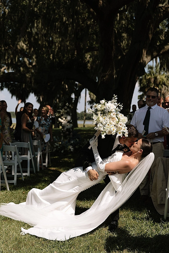 Wedding kiss portrait of bride in strapless gown and veil dipped by groom in suit, holding large white bouquet on tree-shaded lawn aisle