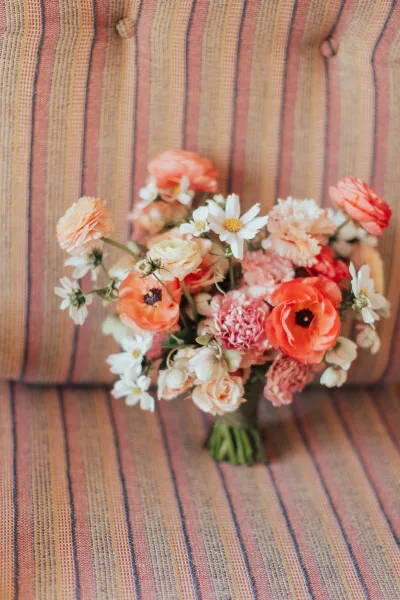 Bridal bouquet wildflower bridal bouquet with coral poppy, ranunculus, daisies, and greenery, resting on a striped upholstered chair