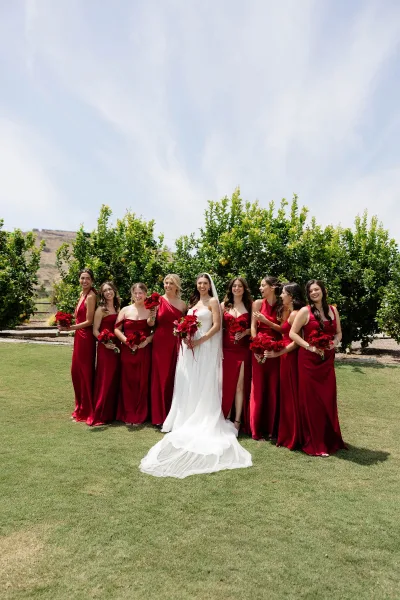 Bridesmaid group photo with bride and bridesmaids in burgundy dresses holding red rose bouquets on a grassy orchard lawn with hills behind