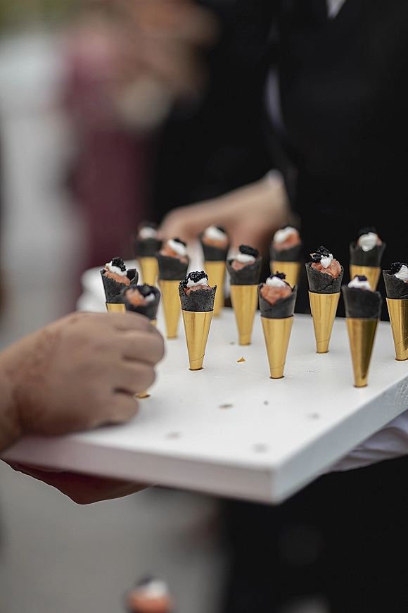 Wedding hors d'oeuvres and passed appetizers wedding served in mini black cones with gold holders on a tray, guests blurred in the background