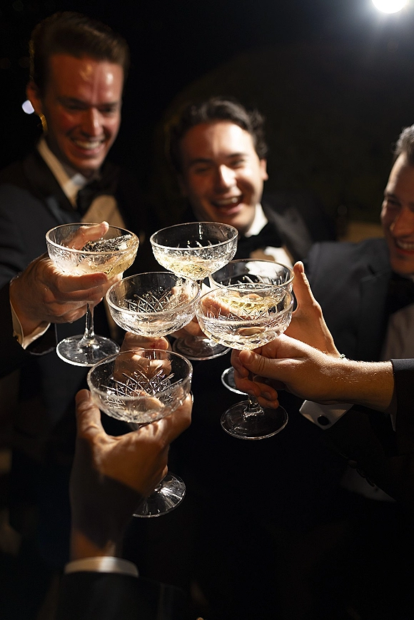 Wedding toast as groomsmen toast in tuxedos clink champagne coupe glasses, cufflinks catching light against dark night bokeh lights
