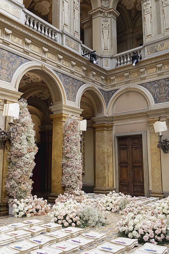 Ceremony setup with indoor ceremony setup featuring blush and white rose floral pillars, aisle floor florals, and cushions beneath ornate arches