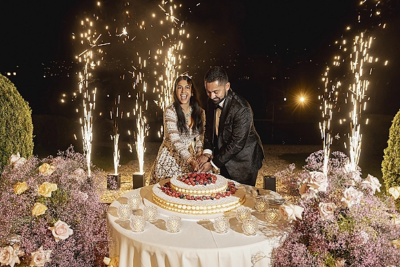 Wedding cake cutting as bride and groom slice a berry-topped two-tier cake with sparklers under string lights on an outdoor lawn at night