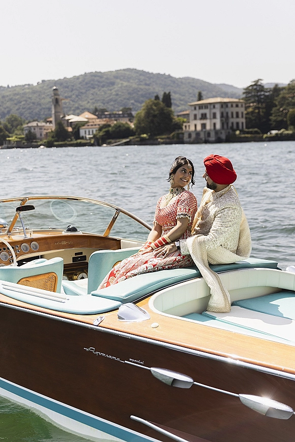 Couple portrait of bride in red lehenga and groom in sherwani with red turban, seated on a boat against a mountain lake backdrop