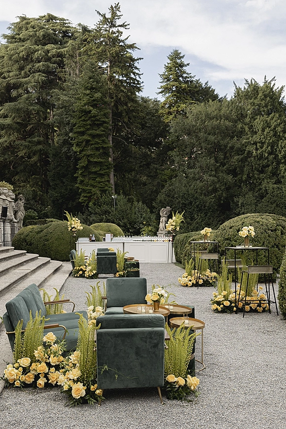 Outdoor lounge decor with velvet chairs, bar stools, and cocktail tables with yellow rose arrangements on a gravel patio by manicured hedges