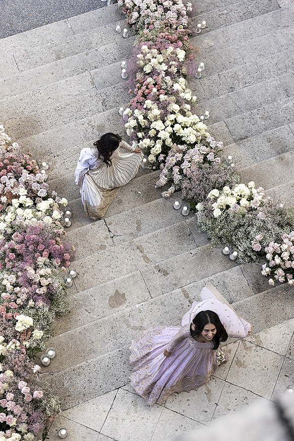 Wedding aisle decor with a floral aisle runner of white and blush roses, baby’s breath, and candles lining a stone staircase outdoors