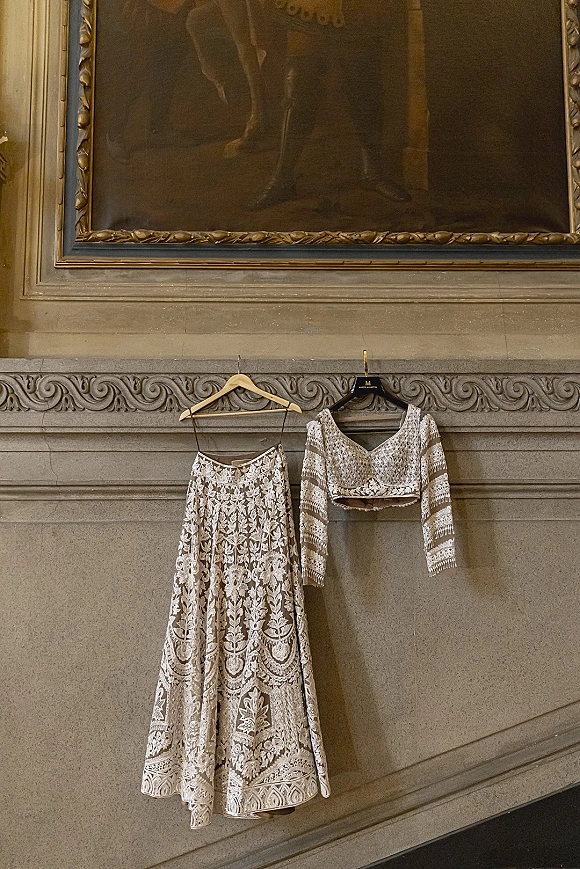 Bridal outfit wedding lehenga hanging on hangers, embroidered skirt and long sleeve blouse against a stone wall with ornate molding