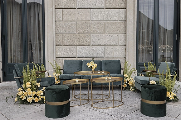 Wedding lounge seating with velvet sofa and chairs around round metal coffee tables, roses and votive candles on a stone patio courtyard