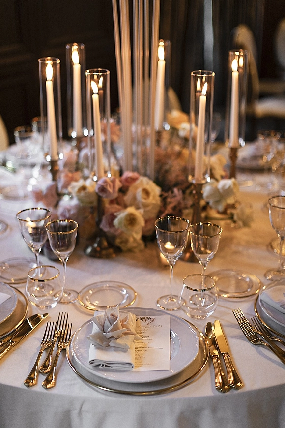 Reception tablescape with wedding table centerpiece, tall taper candles and hurricane holders, blush florals, gold flatware, and menu cards on white linens