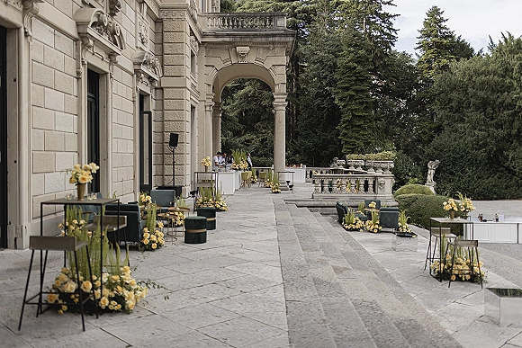 Cocktail hour lounge with green velvet chairs, yellow floral arrangements, and candlelit cocktail tables on a stone terrace by an arched doorway