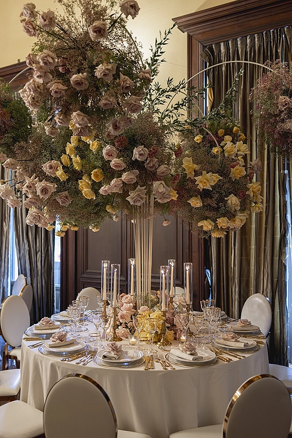 Reception tablescape with a tall floral centerpiece of roses and yellow blooms, taper candles, and gold flatware beneath a hanging floral installation
