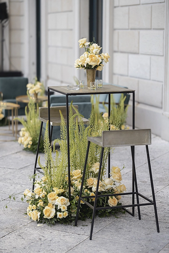 Cocktail table decor with yellow rose arrangement and greenery in bud vases, glass votive candles on a tall table on a stone patio