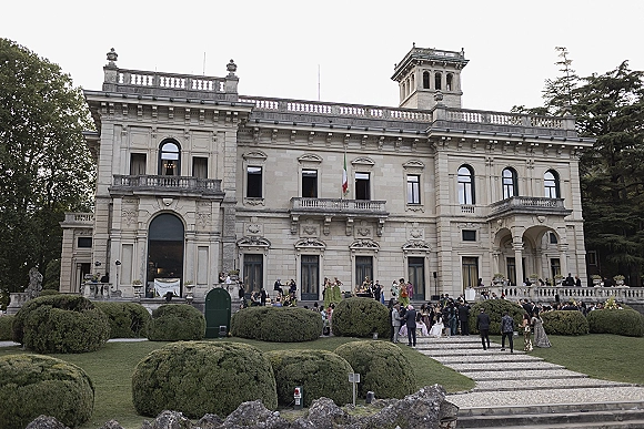 Wedding venue exterior with stone balustrades and arched windows, columns and terrace overlooking manicured hedges and a gravel walkway.