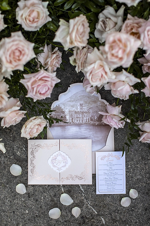 Invitation suite wedding invitation flat lay with blush roses, greenery, rose petals, and ornate cards arranged on a stone surface