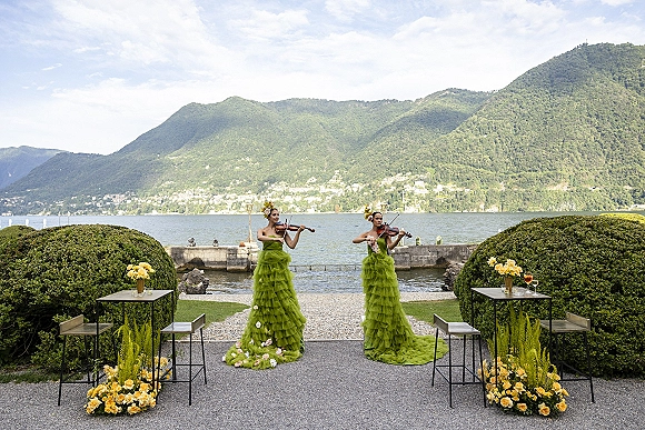 Wedding musicians, violinists at wedding in green tulle gowns play on a lakeside terrace with yellow roses, mountains and cocktail tables behind