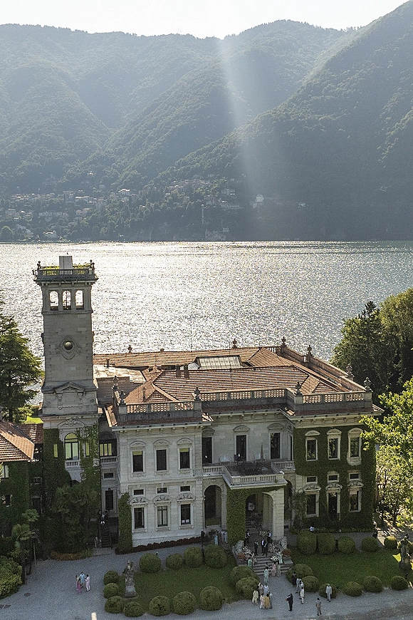 Wedding venue exterior with a historic villa and clock tower, ivy-covered facade and terracotta roof overlooking a sunlit lake and mountains