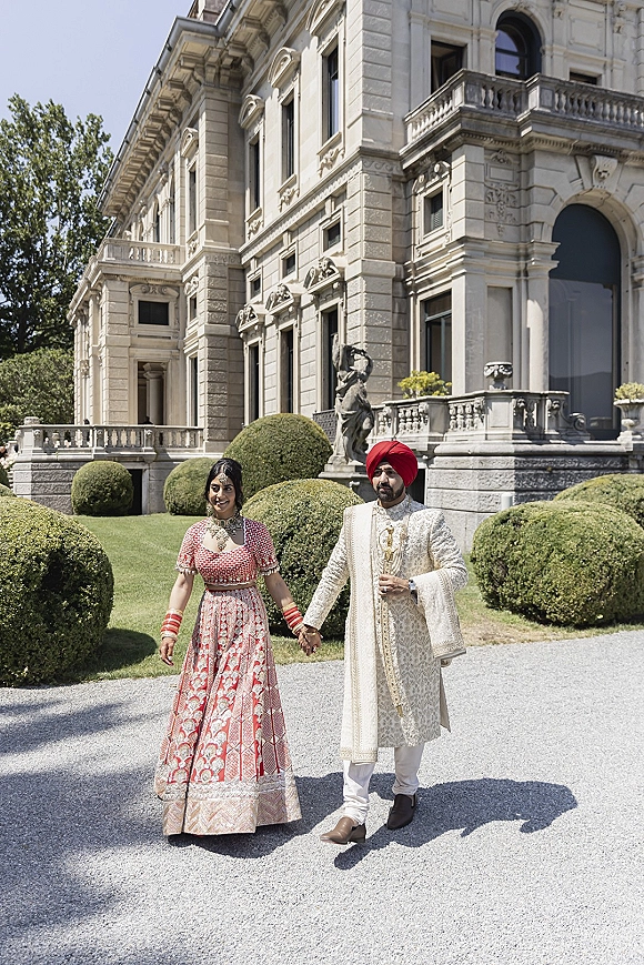Couple portrait of an indian wedding couple holding hands, bride in red lehenga and groom in ivory sherwani before a stone mansion garden backdrop